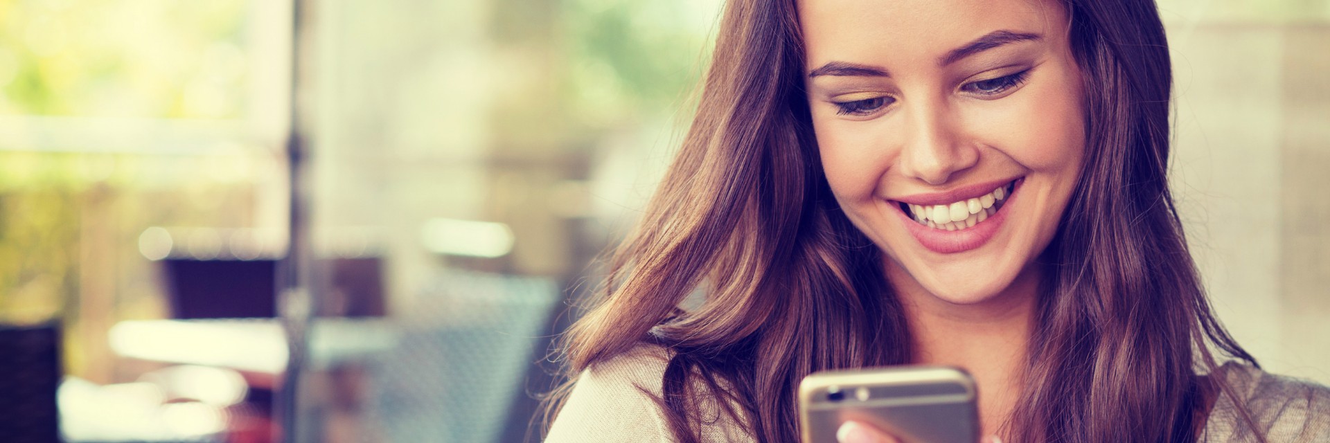 Smiling young woman using smartphone in a bright setting with blurred background.