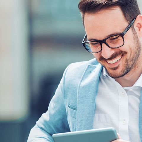 Man in a light blue blazer smiles while using a tablet in a modern setting.