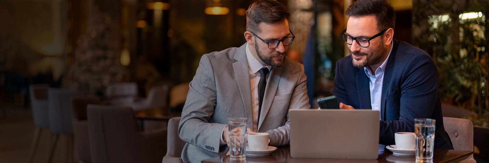 Two men in business attire discussing work over a laptop at a café table with coffee and water glass