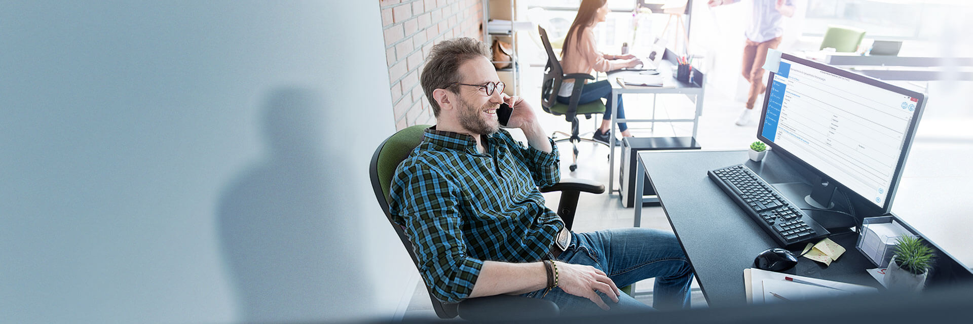 Man sitting at a desk on a phone call, with a computer monitor and other workers visible in the back