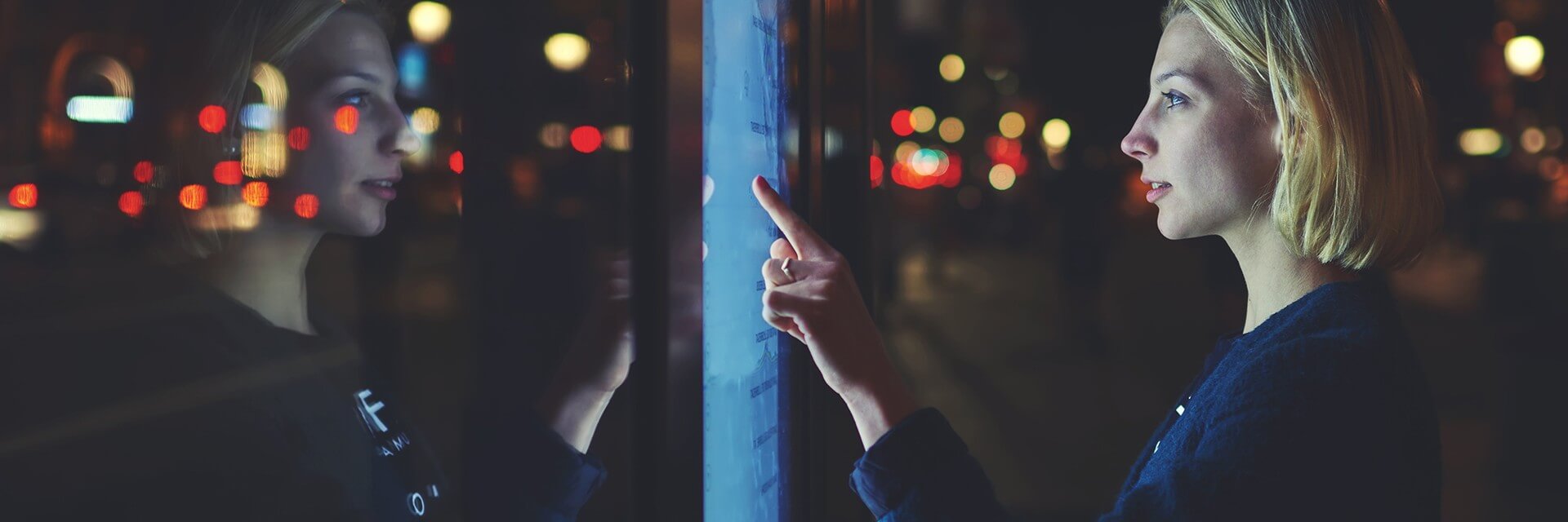 Woman with short blonde hair interacting with a touchscreen in a dimly lit urban setting, with color