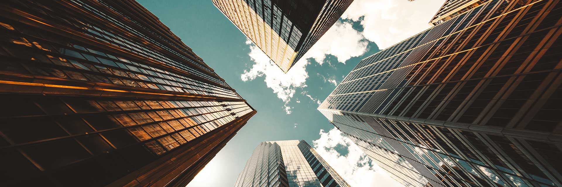 Skyscrapers viewed from below against a blue sky with clouds, highlighting reflections on glass faça