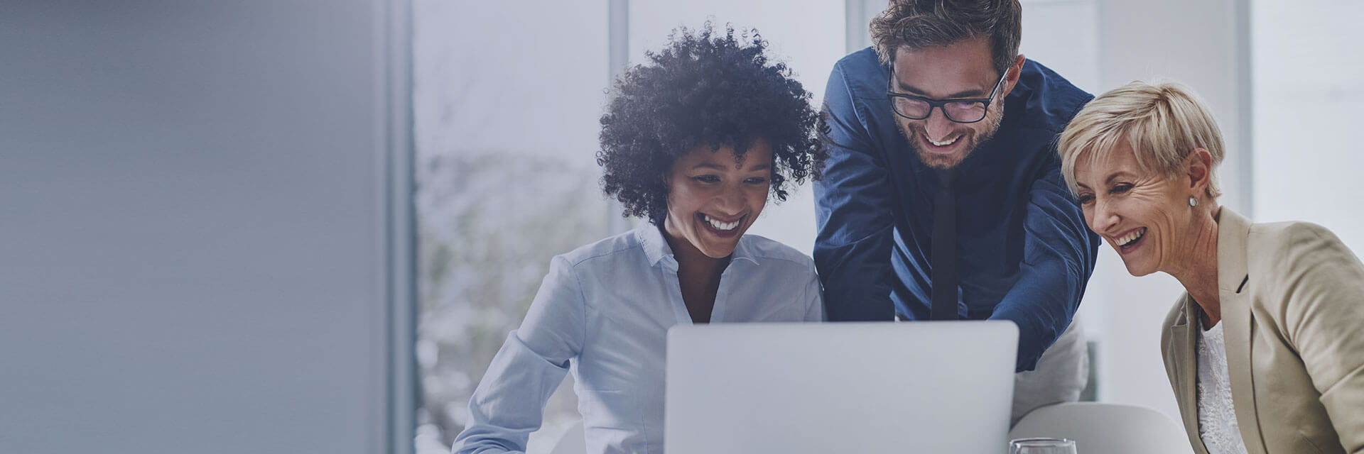 Three colleagues smiling and discussing work while looking at a laptop in a bright office setting.